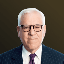 Elderly man with white hair and glasses, wearing a dark pinstripe suit, white shirt, and purple tie, against a plain background.
